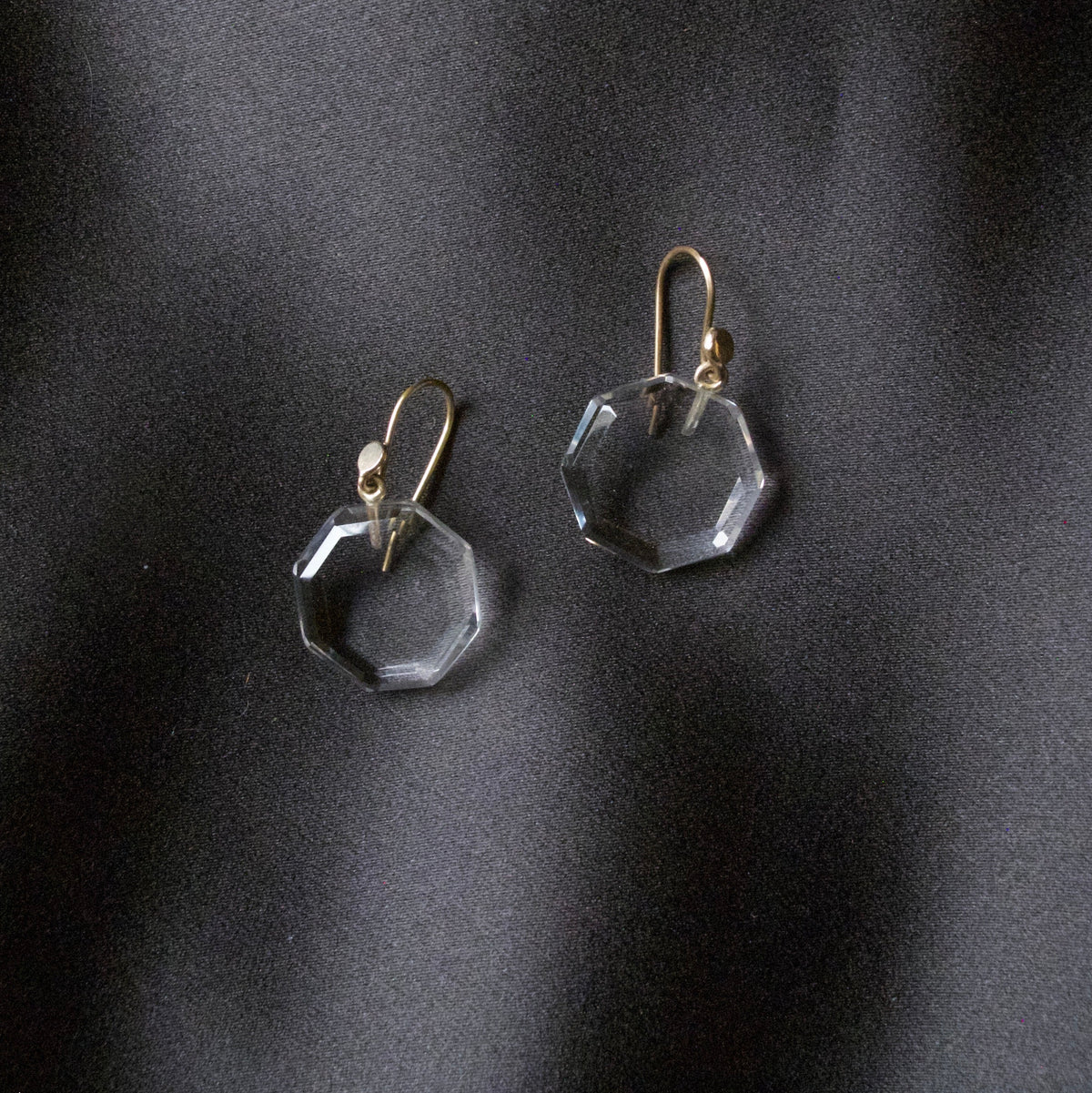 A pair of clear faceted octagonal drop earrings with gold fishhook wires, displayed on a dark, textured satin fabric background.