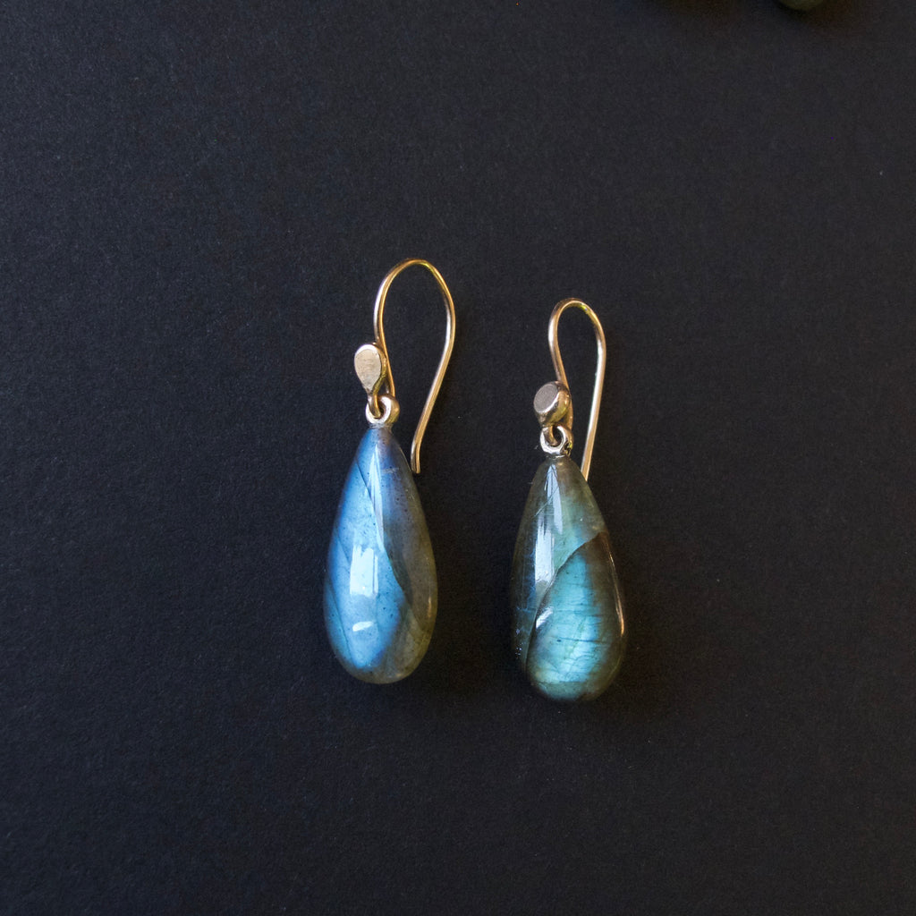 A pair of teardrop-shaped labradorite drop earrings with gold fishhook wires, displayed on a dark textured background.