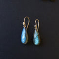 A pair of teardrop-shaped labradorite drop earrings with gold fishhook wires, displayed on a dark textured background.