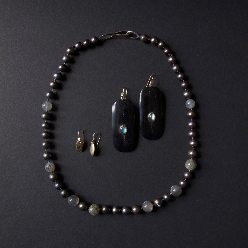 A jewelry set featuring a beaded necklace and matching earrings arranged on a dark background. The necklace and pendants are made of dark black stone with iridescent blue-green inclusions, accented with small gray beads and gold-toned hooks.