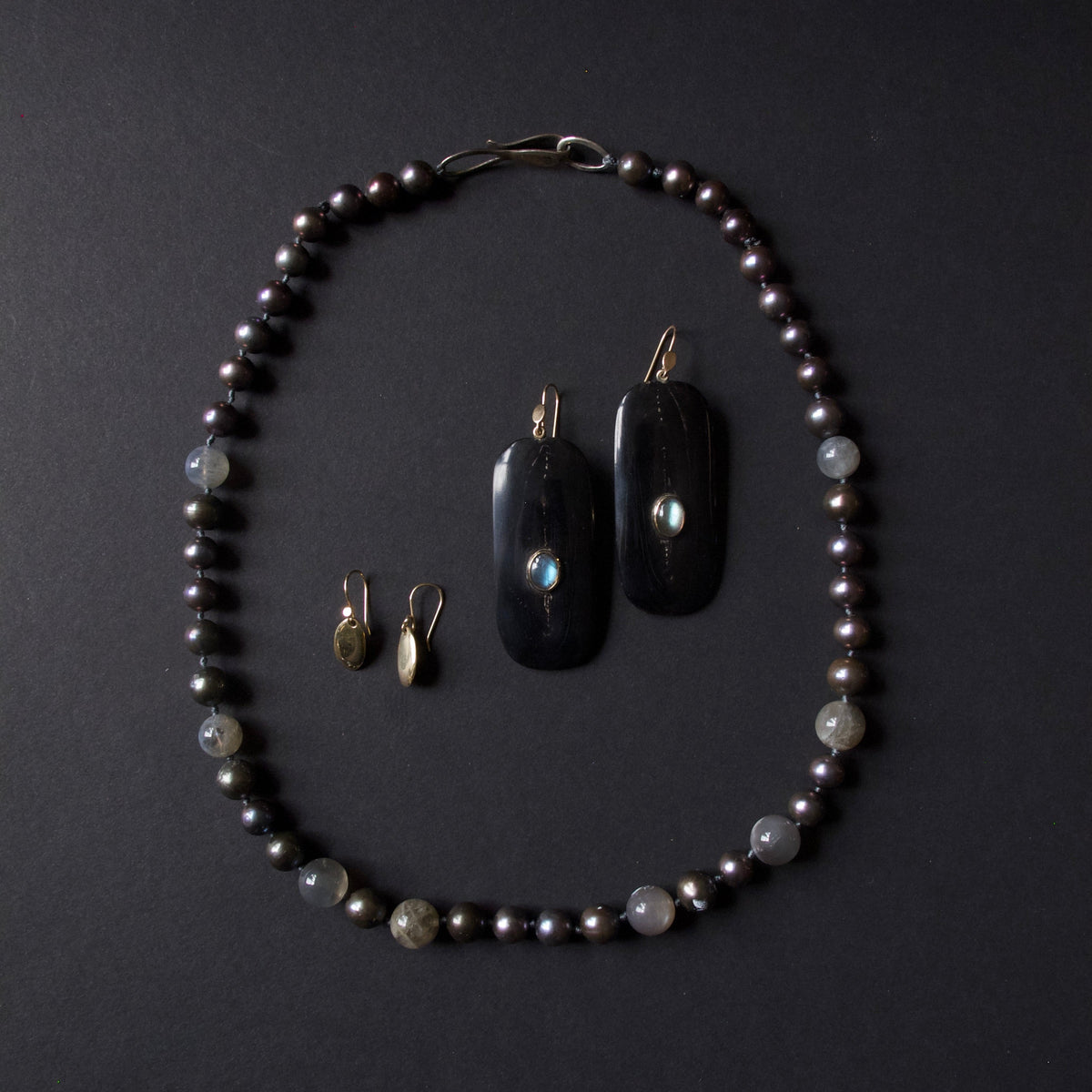 A jewelry set featuring a beaded necklace and matching earrings arranged on a dark background. The necklace and pendants are made of dark black stone with iridescent blue-green inclusions, accented with small gray beads and gold-toned hooks.