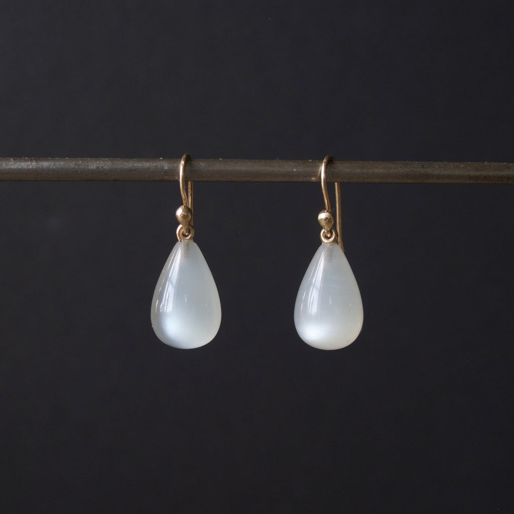 A pair of teardrop-shaped white stone drop earrings with gold hooks, hanging from a dark horizontal bar against a black background.