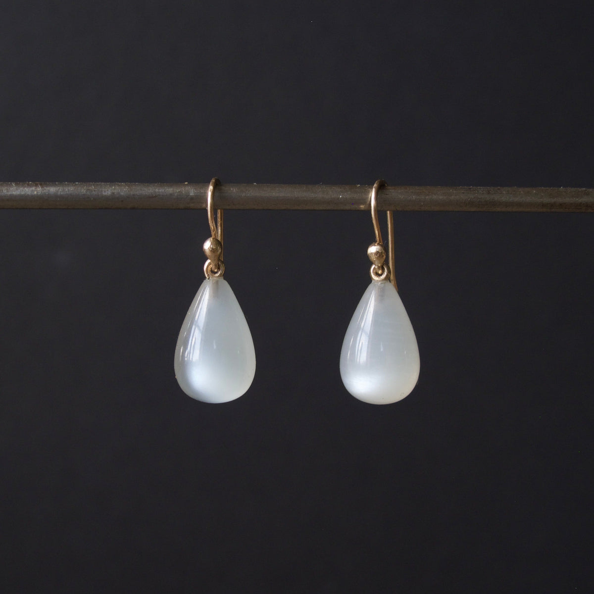 A pair of teardrop-shaped white stone drop earrings with gold hooks, hanging from a dark horizontal bar against a black background.