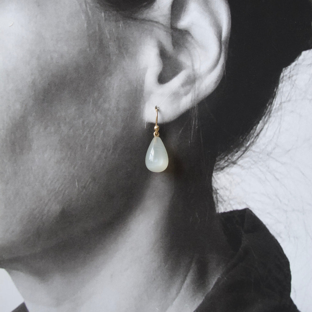 A close-up side profile of a woman's ear wearing a small gold hoop earring with a teardrop-shaped milky white stone drop.