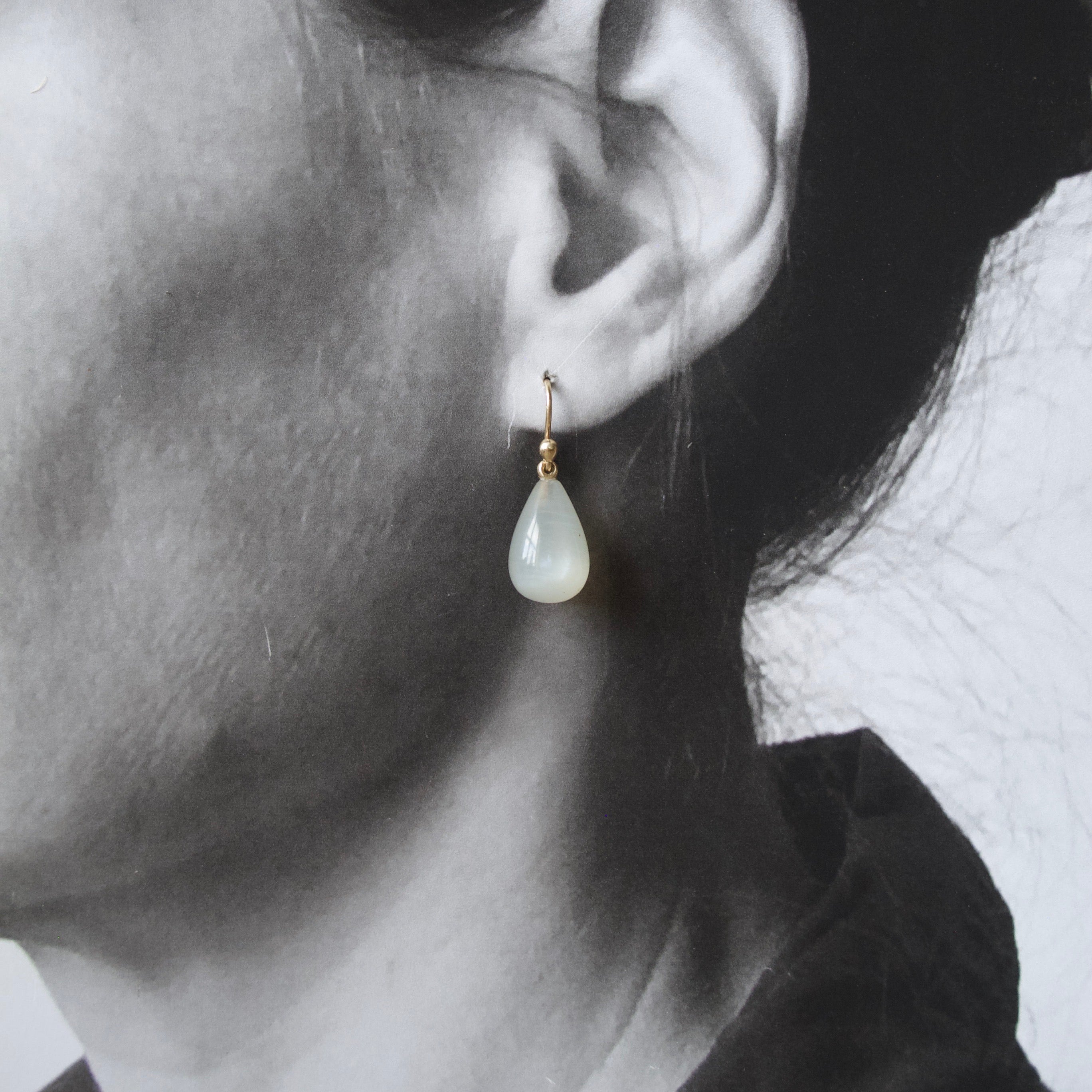 A close-up side profile of a woman's ear wearing a small gold hoop earring with a teardrop-shaped milky white stone drop.
