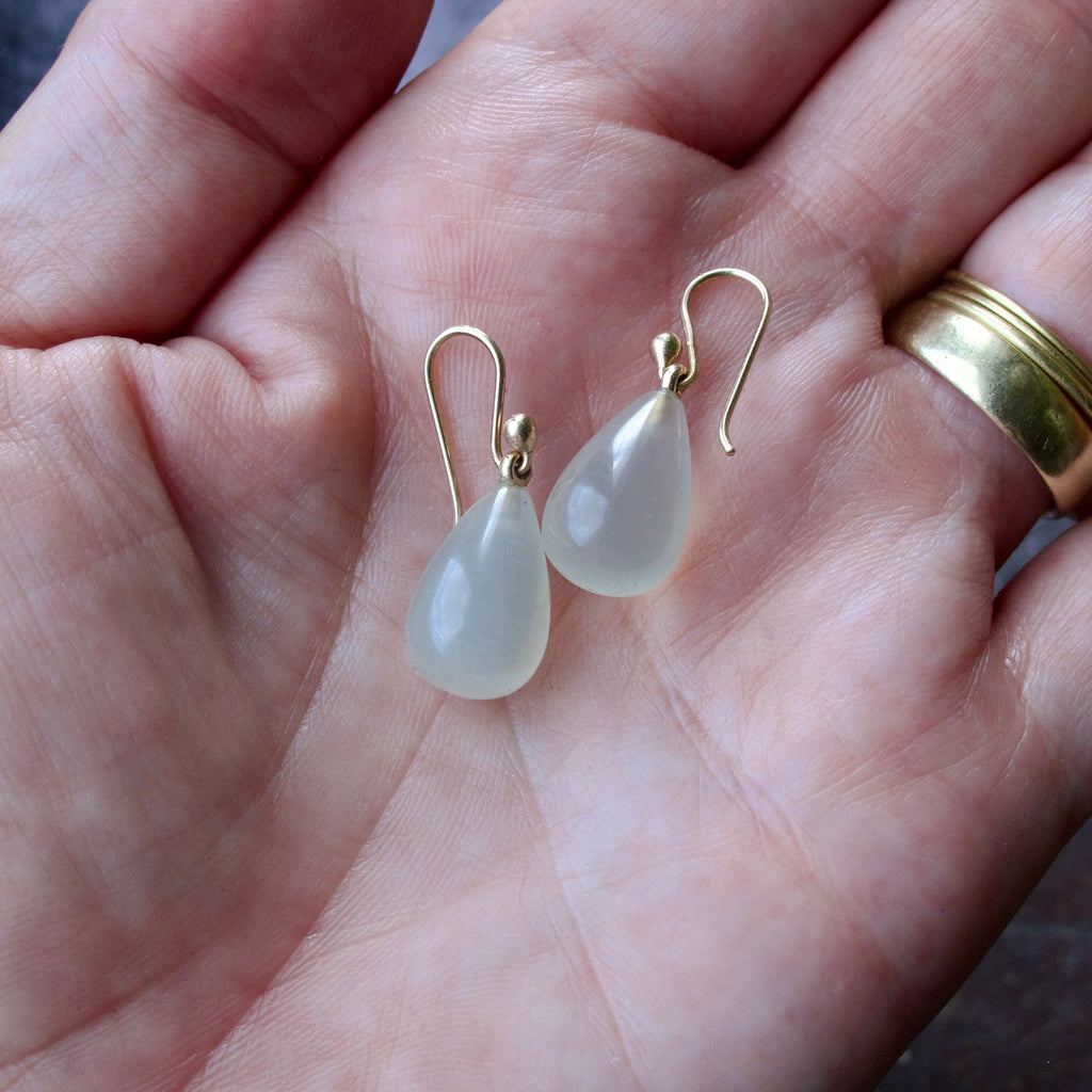 A pair of gold-tone drop earrings with teardrop-shaped milky white stones, shown resting on an open hand. The hand also features a gold band ring on the ring finger.