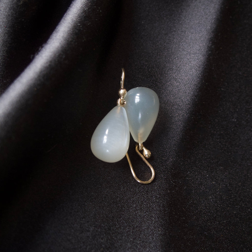 A pair of gold drop earrings with teardrop-shaped milky white beads, displayed on a draped black satin fabric.