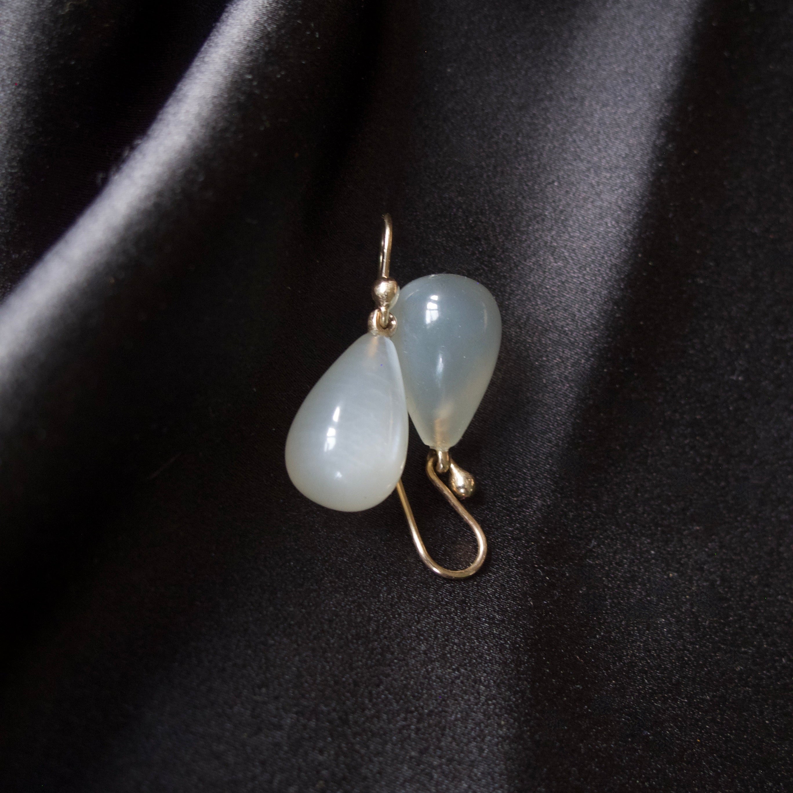 A pair of gold drop earrings with teardrop-shaped milky white beads, displayed on a draped black satin fabric.