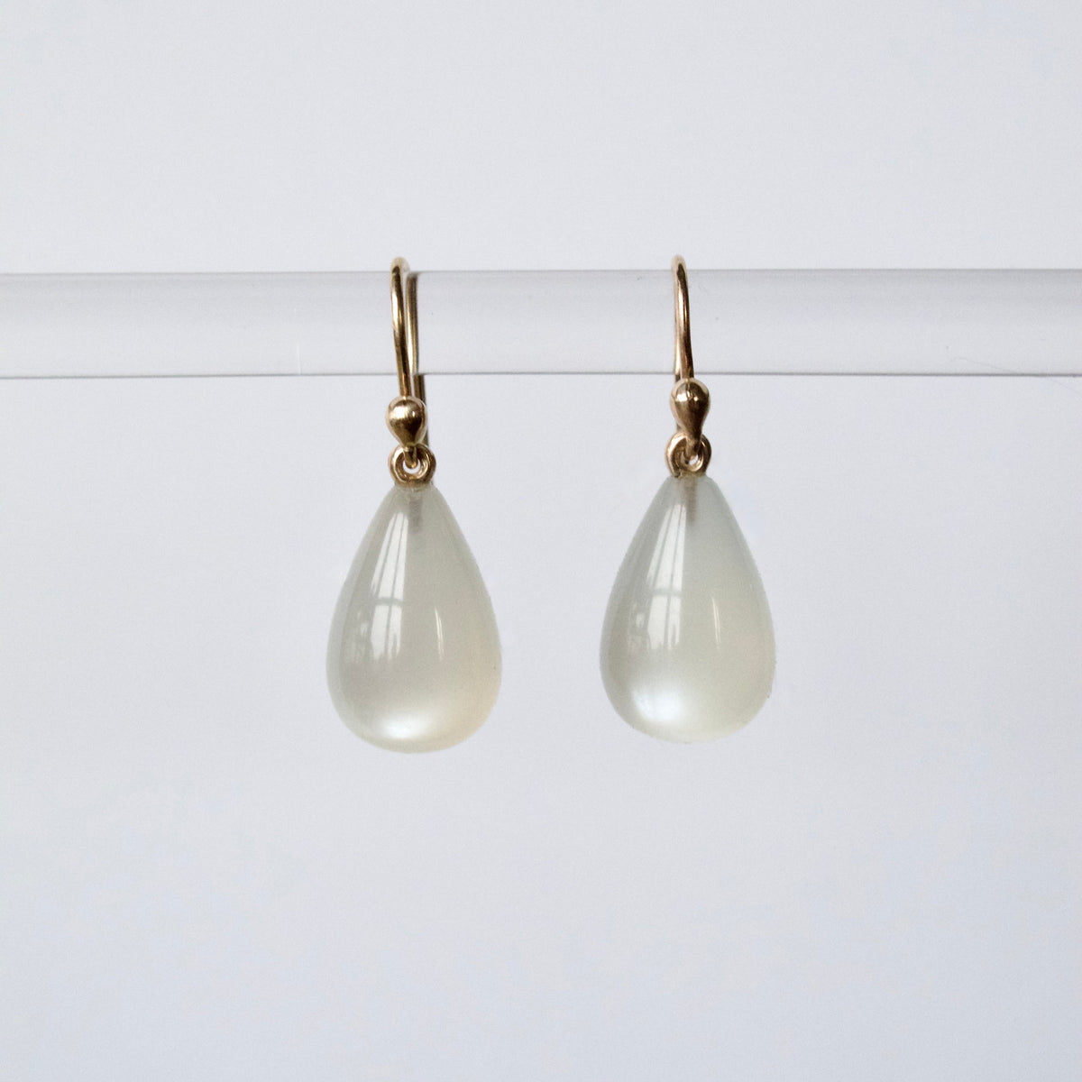 A pair of teardrop-shaped drop earrings with milky white stone pendants and gold hooks, hanging from a thin horizontal bar against a light gray background.