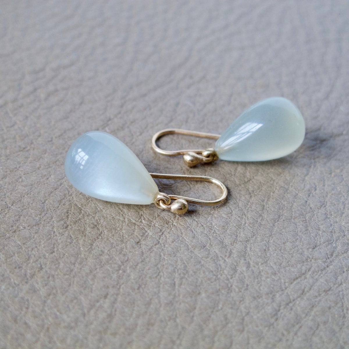 A pair of gold drop earrings with teardrop-shaped milky white cabochon gemstones, displayed on a textured beige surface.