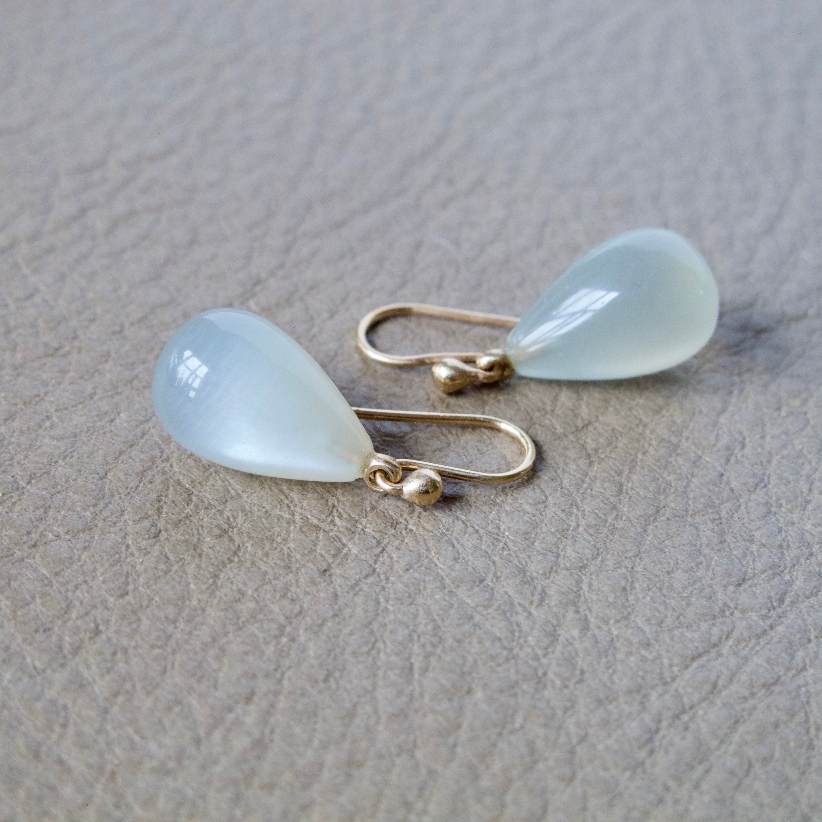 A pair of gold drop earrings with teardrop-shaped milky white cabochon gemstones, displayed on a textured beige surface.