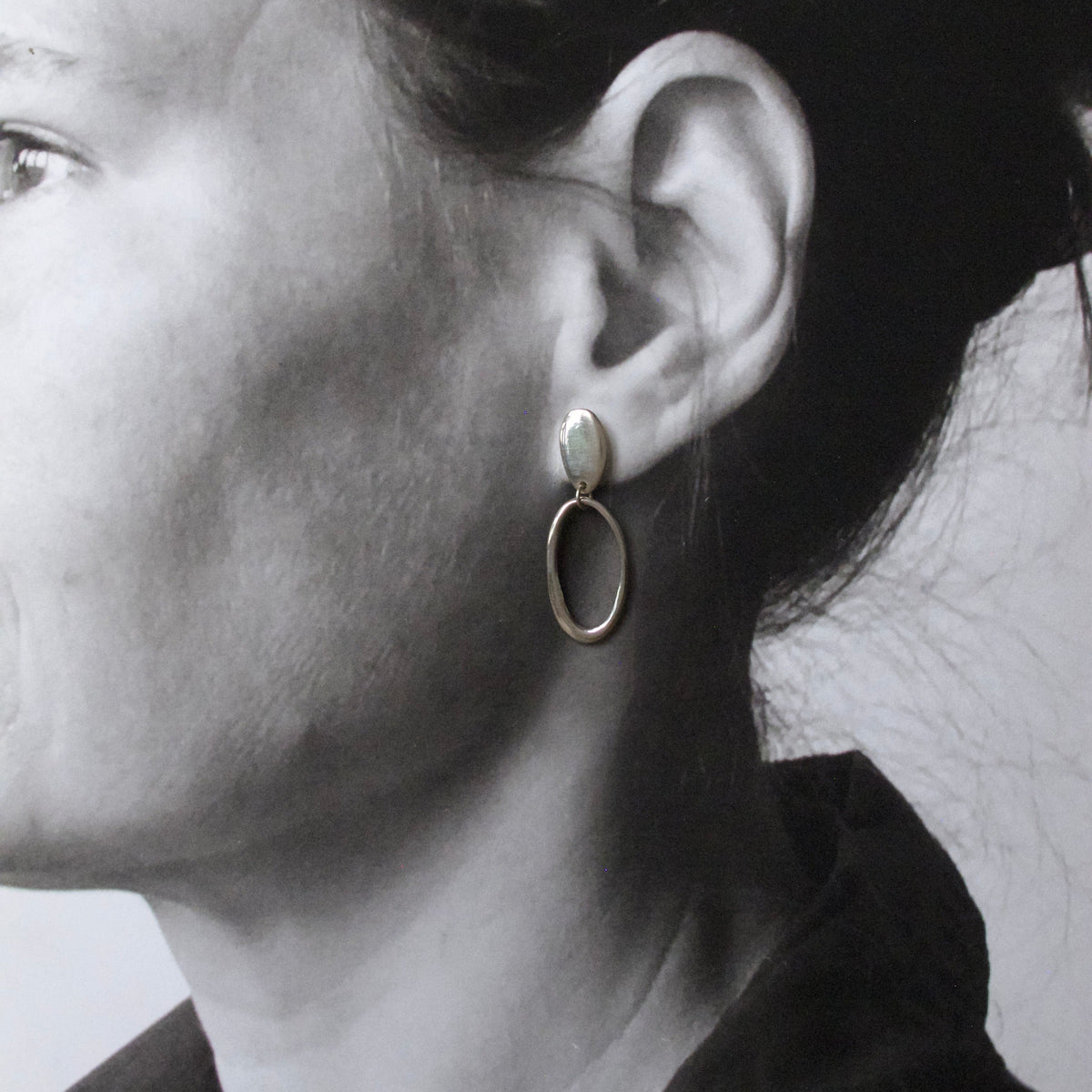 A close-up side profile of a woman wearing a gold-toned drop earring with a small oval stone and a larger hanging oval loop.