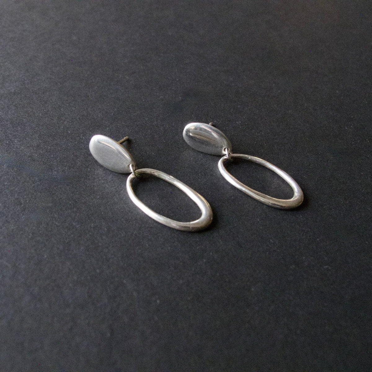 A pair of minimalist silver drop earrings with flat oval disc tops and open oval hoops below, displayed on a dark textured surface.