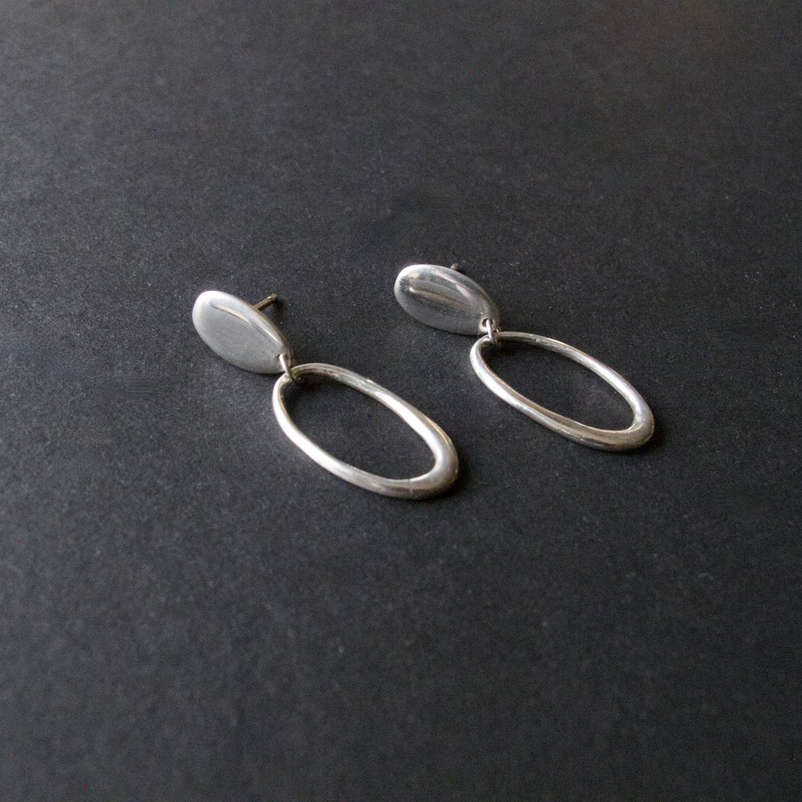 A pair of minimalist silver drop earrings with flat oval disc tops and open oval hoops below, displayed on a dark textured surface.
