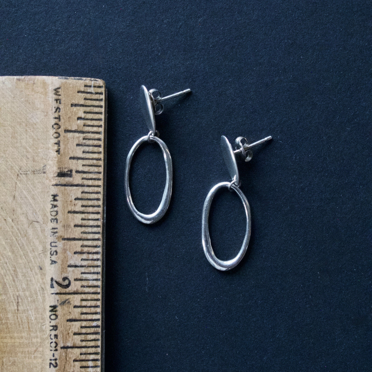 A pair of silver oval drop earrings displayed next to a wooden ruler on a dark navy blue background.