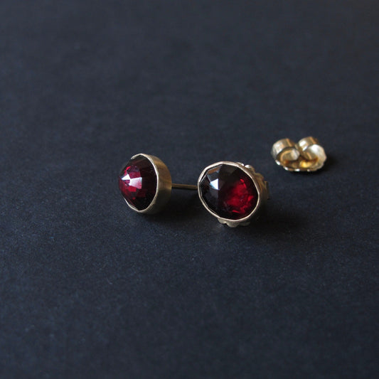 A pair of gold stud earrings with faceted dark red gemstones, shown on a dark textured background with one earring's backing visible.