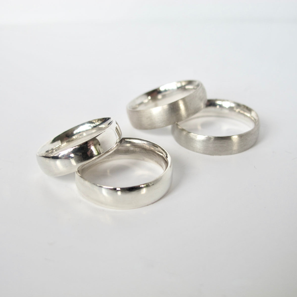 Four silver bands, including two matte textured rings and two polished smooth rings, arranged on a plain white background.