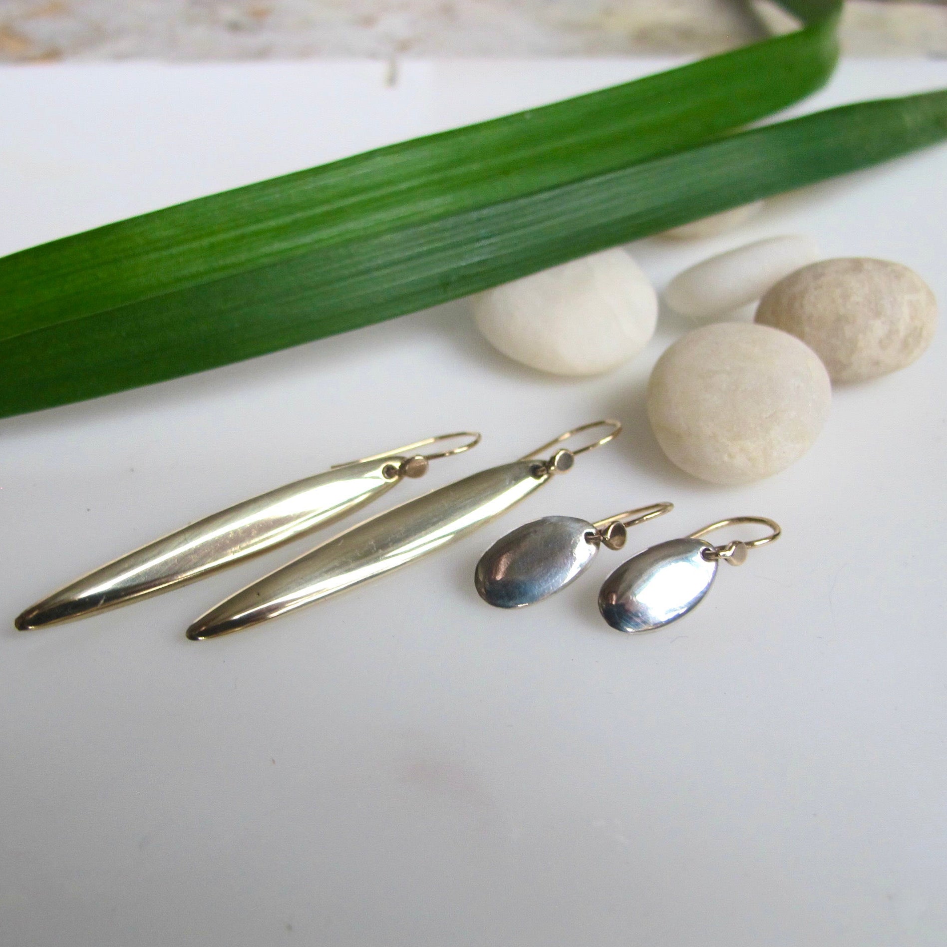 Two gold-toned elongated drop earrings and two silver-toned oval drop earrings arranged on a white surface with smooth stones and a green leaf in the background.