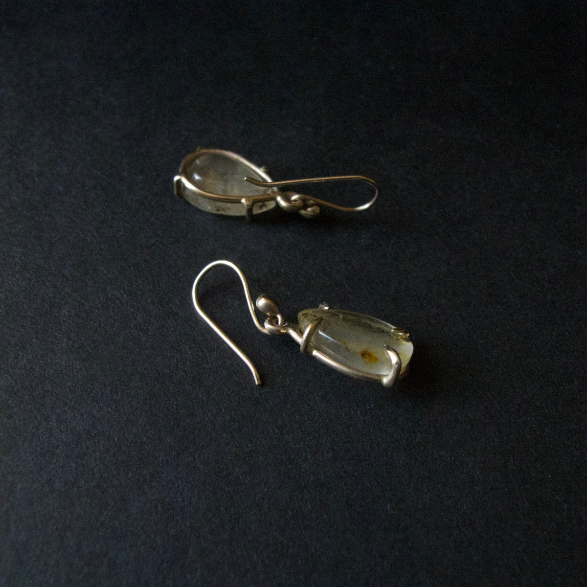 A pair of handmade dangle earrings featuring small, irregular pale greenish-gray stones wrapped in silver wire, suspended from silver fishhook ear wires, displayed on a dark textured background.