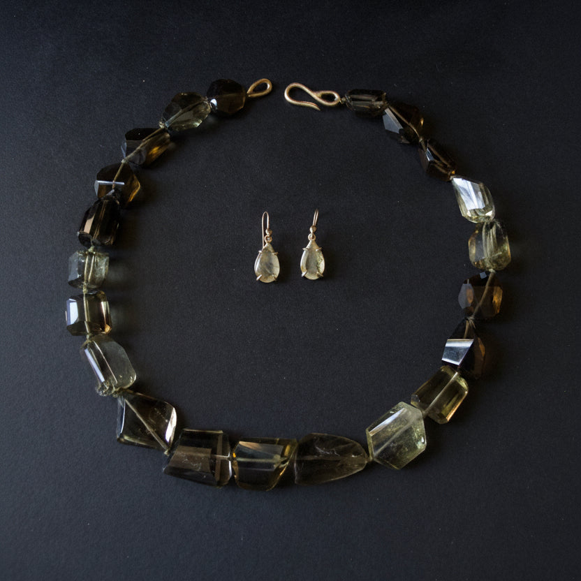 A beaded necklace and matching teardrop dangle earrings laid flat on a dark background. The jewelry features faceted translucent smoky amber and olive-green beads with gold-tone metal findings.