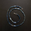 A beaded necklace made of elongated, polished blue beads with silver-toned metal clasp and ring, arranged in a spiral on a dark background.