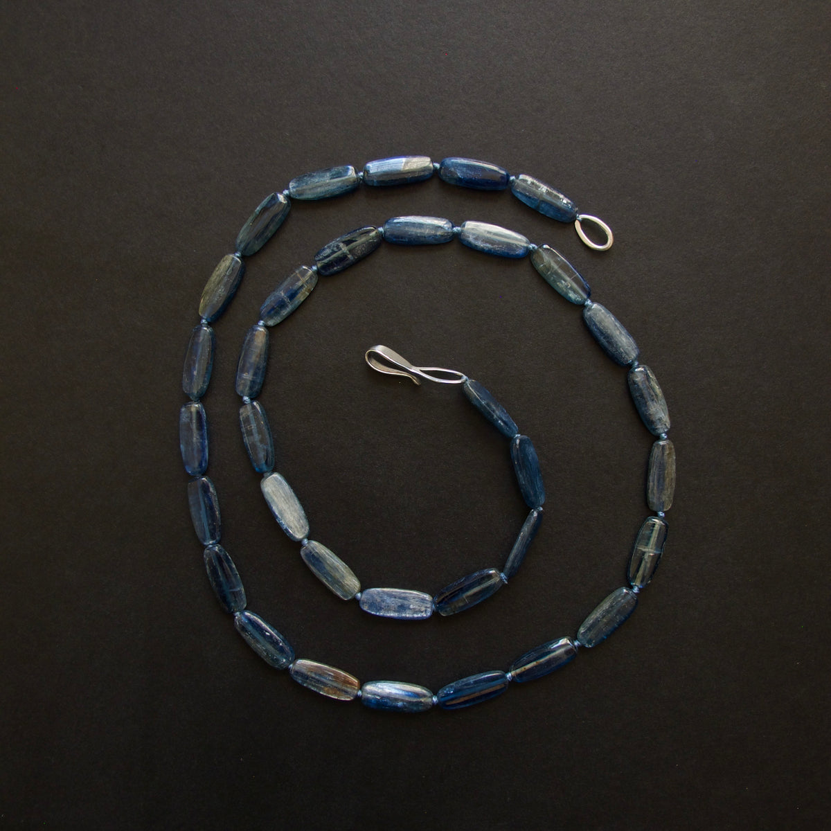 A beaded necklace made of elongated, polished blue beads with silver-toned metal clasp and ring, arranged in a spiral on a dark background.