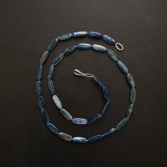 A beaded necklace made of elongated, polished blue beads with silver-toned metal clasp and ring, arranged in a spiral on a dark background.