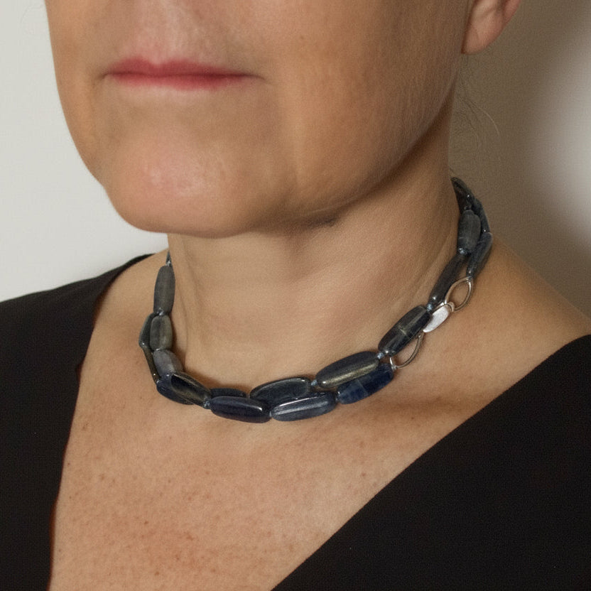 A close-up of a woman wearing a chunky dark blue-gray beaded necklace with silver-toned metal links, paired with a black V-neck top.