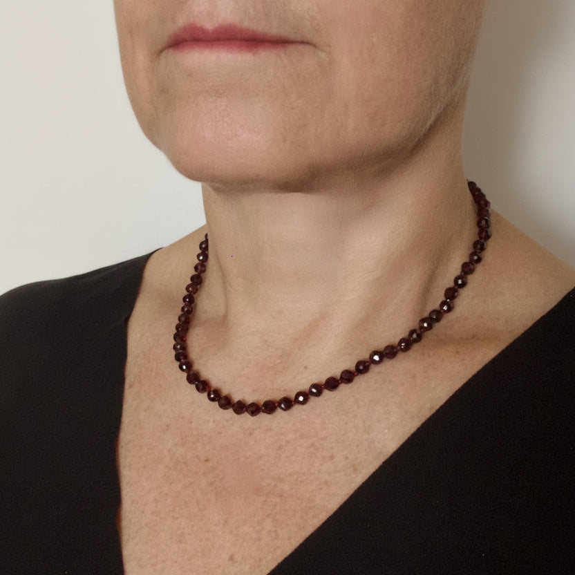 A close-up of a woman wearing a dark reddish-brown beaded necklace, styled with a black V-neck top against a light textured wall.