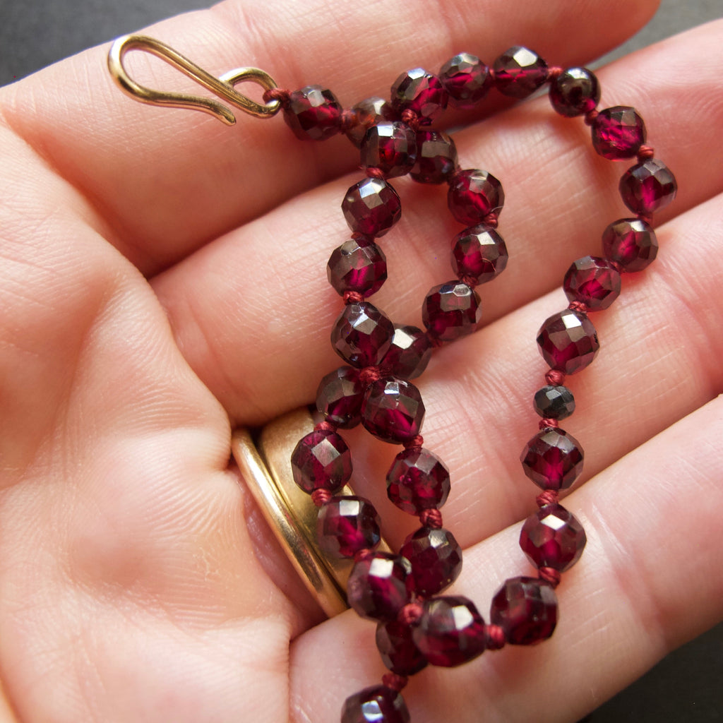A close-up of a hand holding a strand of faceted dark red beads with a gold-tone lobster clasp, with a gold ring on the finger.