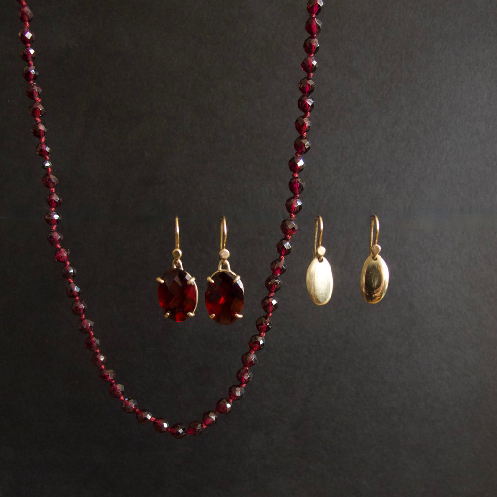 A jewelry set displayed on a dark background, featuring a long beaded necklace in deep red, a pair of red drop earrings with gold hooks, and a pair of larger oval gold earrings with cream inlays.