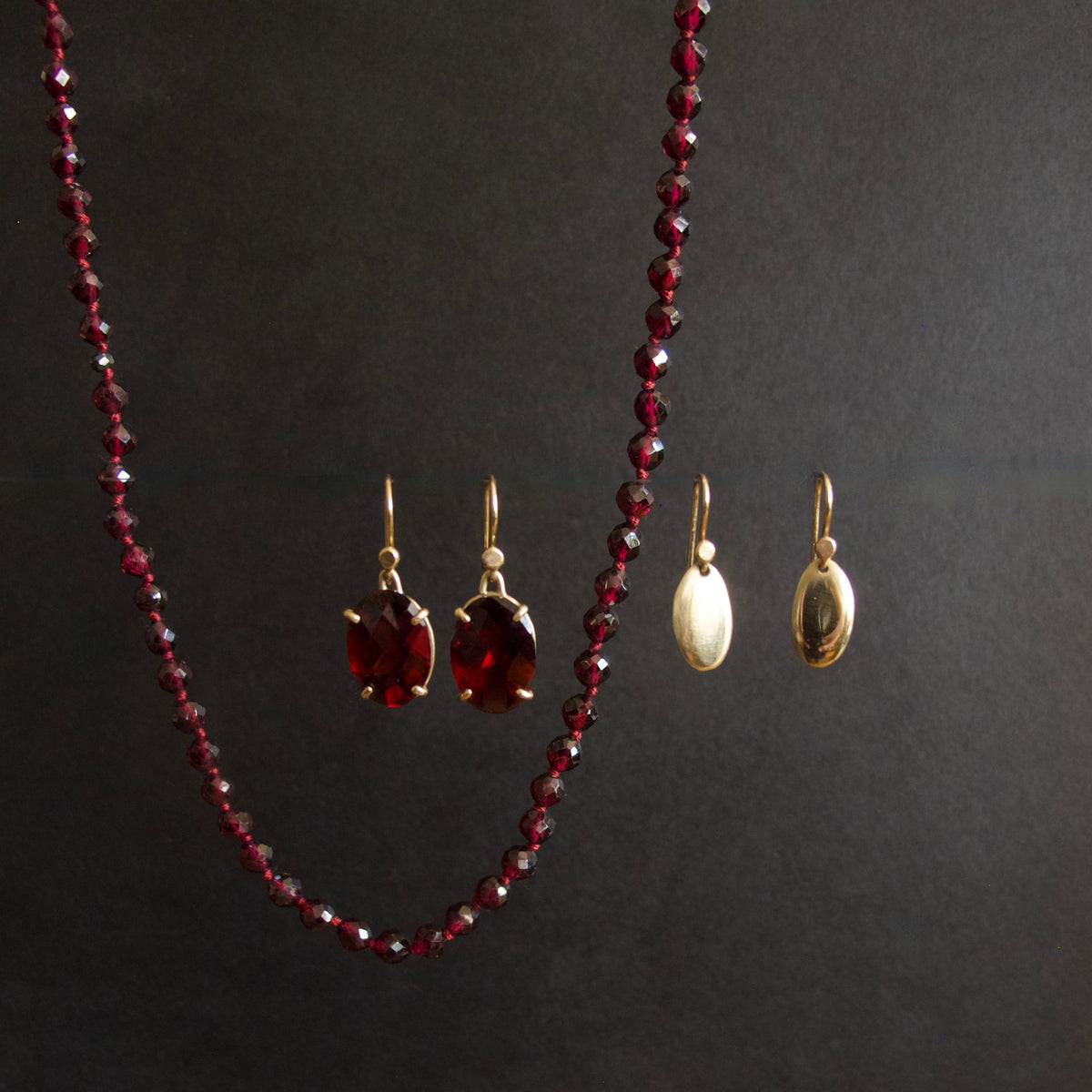 A jewelry set displayed on a dark background, featuring a long beaded necklace in deep red, a pair of red drop earrings with gold hooks, and a pair of larger oval gold earrings with cream inlays.