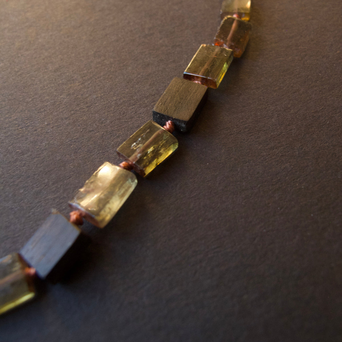 A close-up of a beaded necklace made of alternating translucent olive-green and dark brown rectangular beads, strung on a dark background.