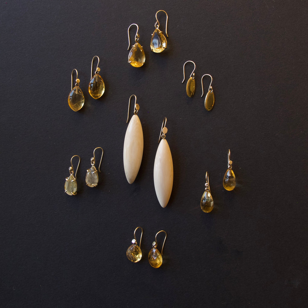 A set of handmade earrings arranged on a dark background, featuring two large cream-colored teardrop beads, several pairs of yellow teardrop gemstone drops, and smaller yellow and green teardrop accents.