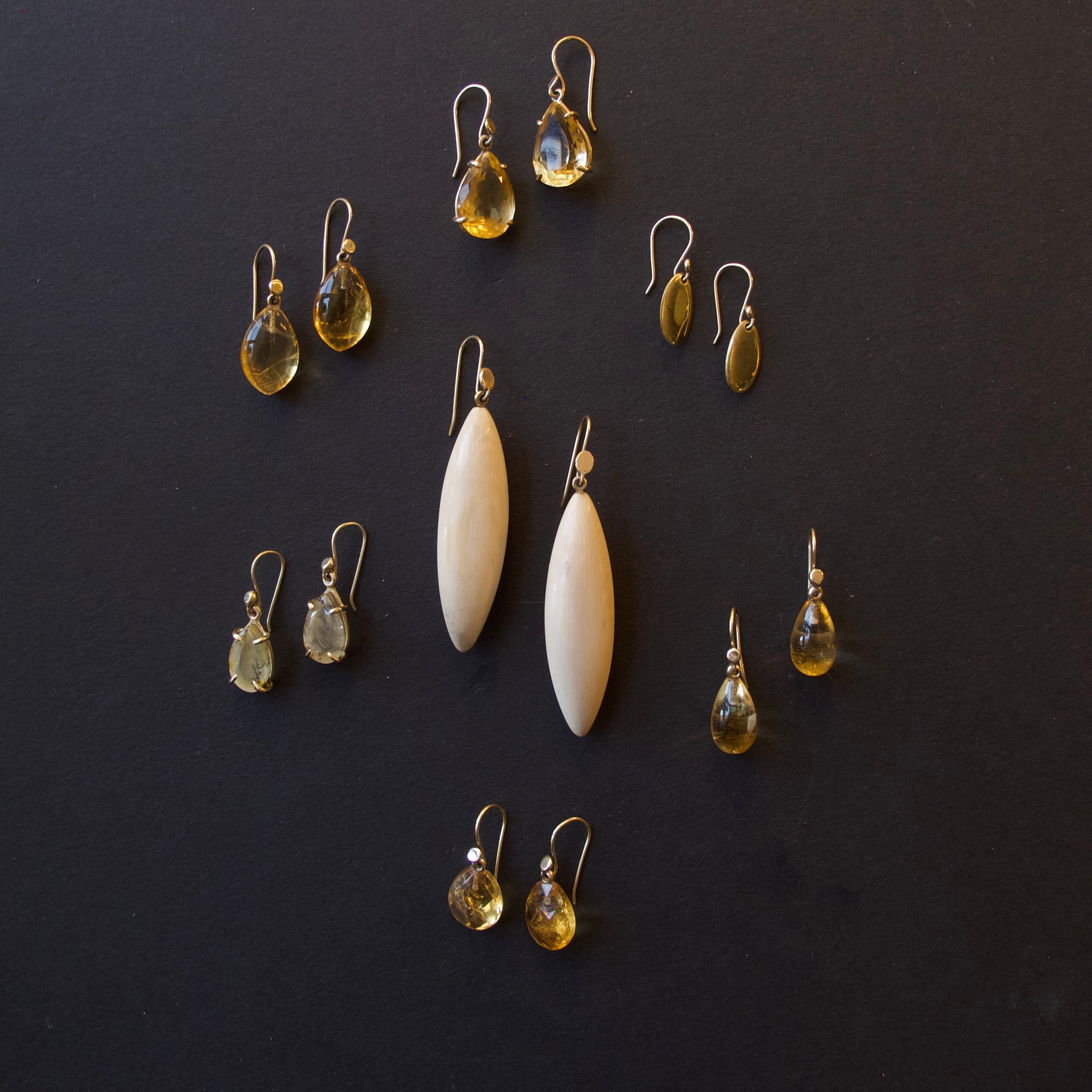 A set of handmade earrings arranged on a dark background, featuring two large cream-colored teardrop beads, several pairs of yellow teardrop gemstone drops, and smaller yellow and green teardrop accents.
