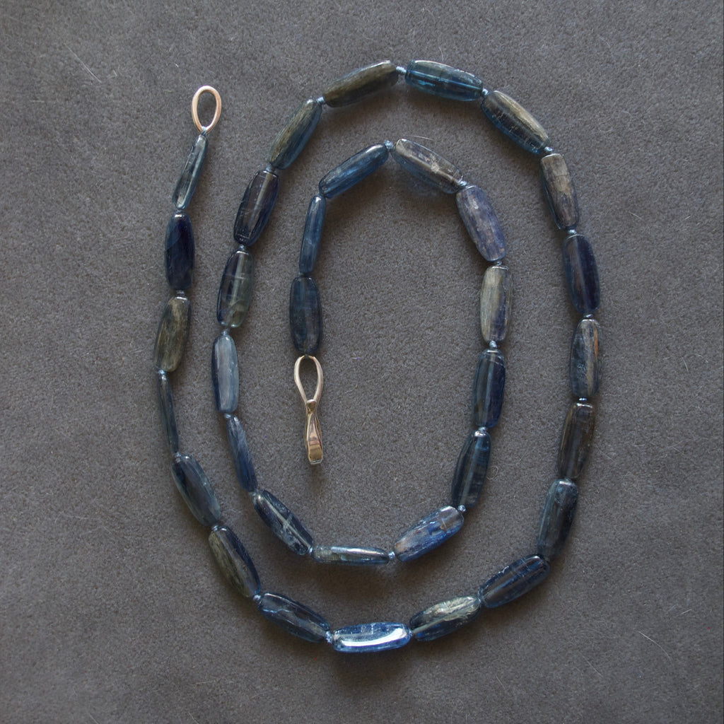 A beaded necklace made of elongated, faceted dark blue beads with a small gold-toned ring clasp, laid out on a dark gray textured background.