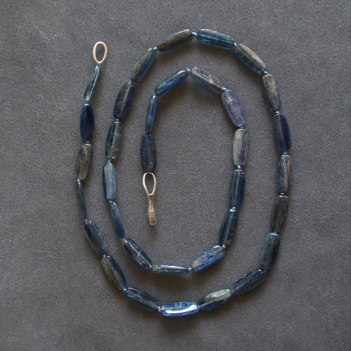A beaded necklace made of elongated, faceted dark blue beads with a small gold-toned ring clasp, laid out on a dark gray textured background.
