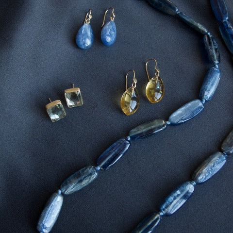 A jewelry flat-lay featuring a long beaded necklace and several pairs of earrings arranged on a dark navy fabric background. The necklace consists of elongated dark blue-gray beads.