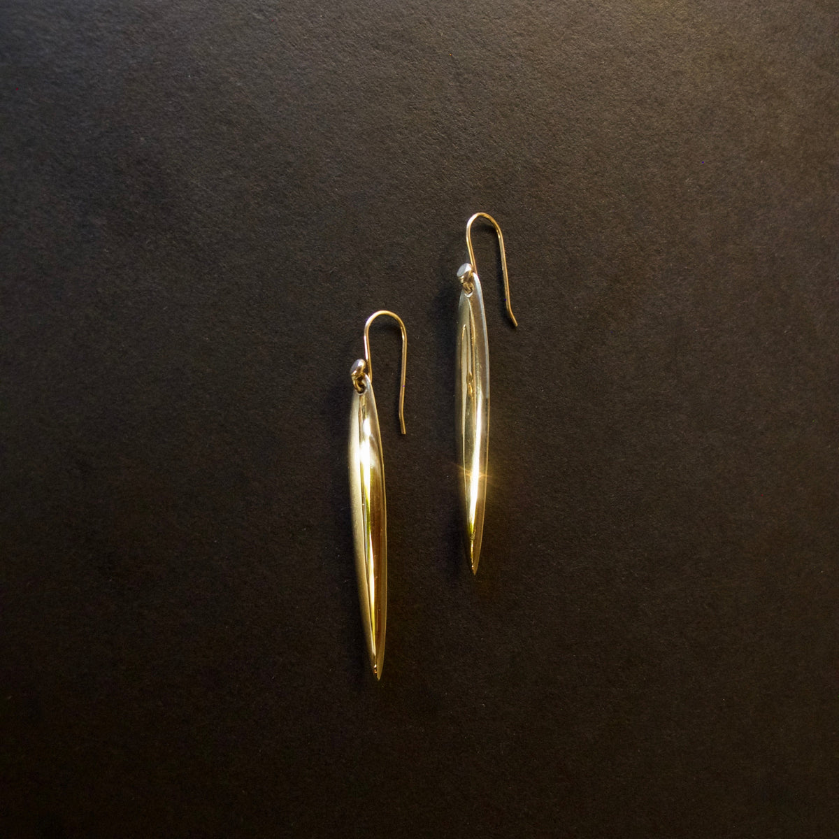 A pair of gold-toned dangle earrings with elongated teardrop-shaped drops and fishhook backs, displayed on a dark textured background.