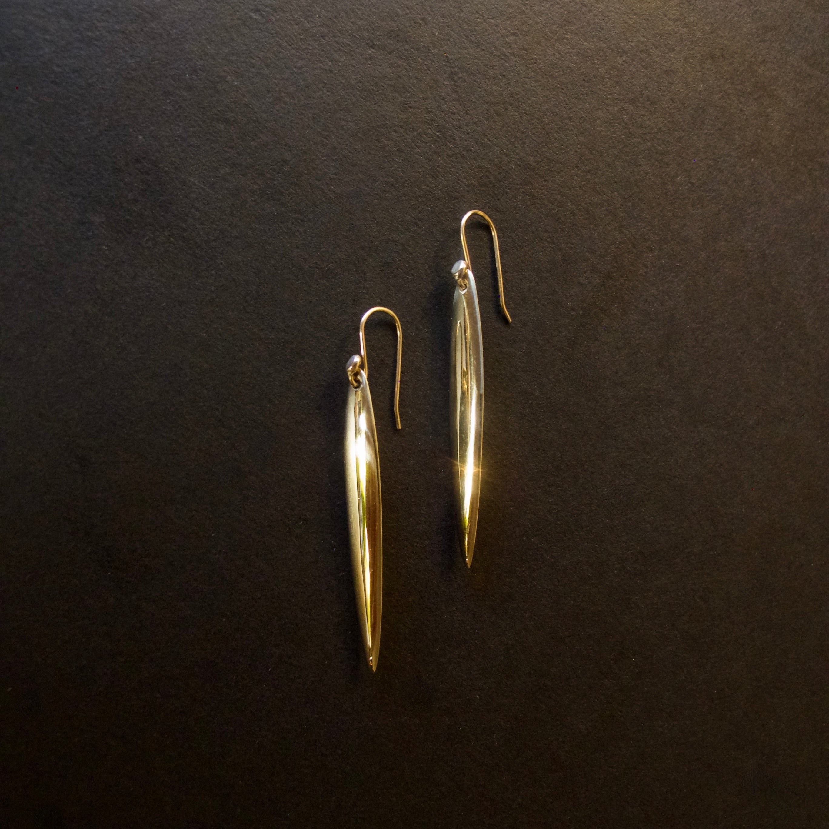 A pair of gold-toned dangle earrings with elongated teardrop-shaped drops and fishhook backs, displayed on a dark textured background.