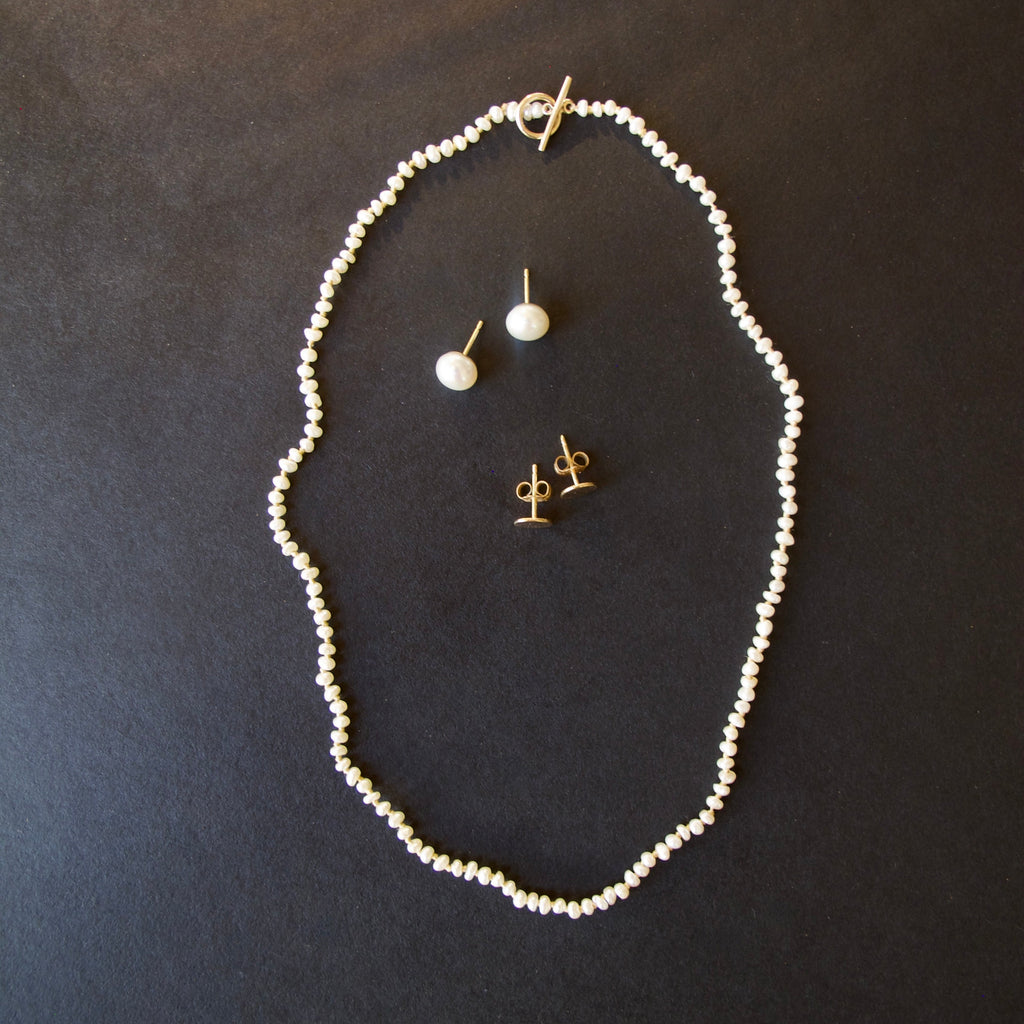 A jewelry set featuring a delicate white pearl necklace, a matching single-drop pearl stud earring, and gold post earrings, arranged on a dark textured background.
