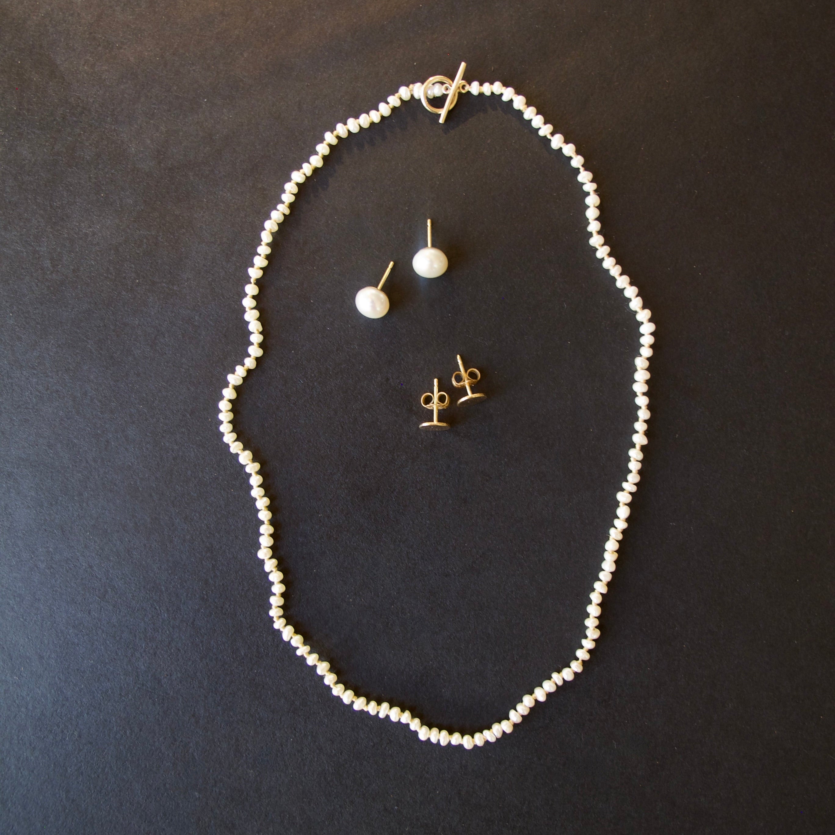 A jewelry set featuring a delicate white pearl necklace, a matching single-drop pearl stud earring, and gold post earrings, arranged on a dark textured background.