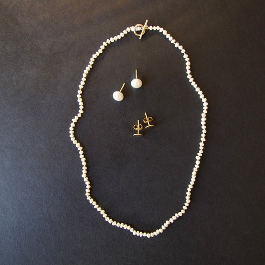 A jewelry set featuring a delicate white pearl necklace, a matching single-drop pearl stud earring, and gold post earrings, arranged on a dark textured background.