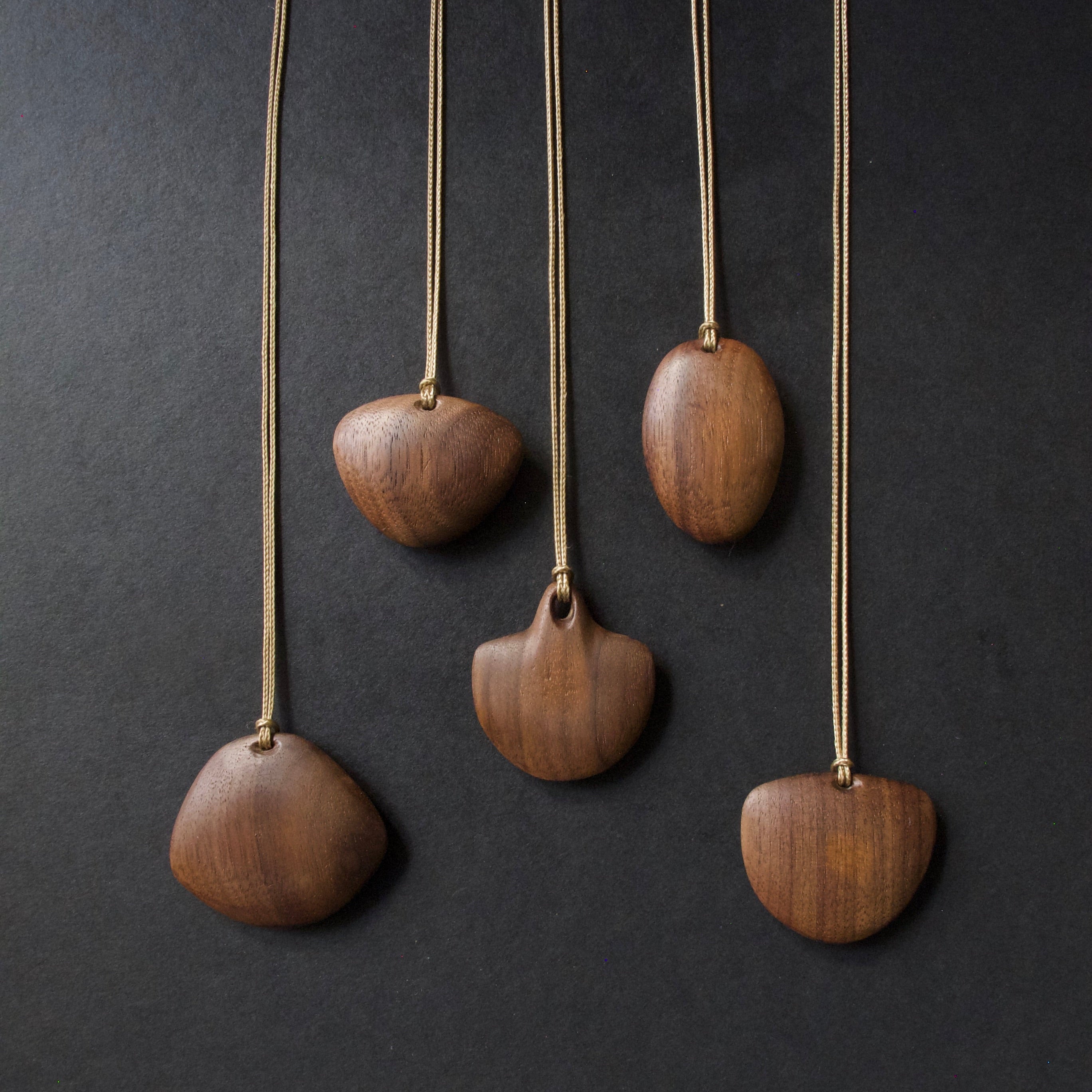 Five wooden pendant necklaces with organic, hand-shaped wood pendants hanging from thin cords against a dark charcoal background.