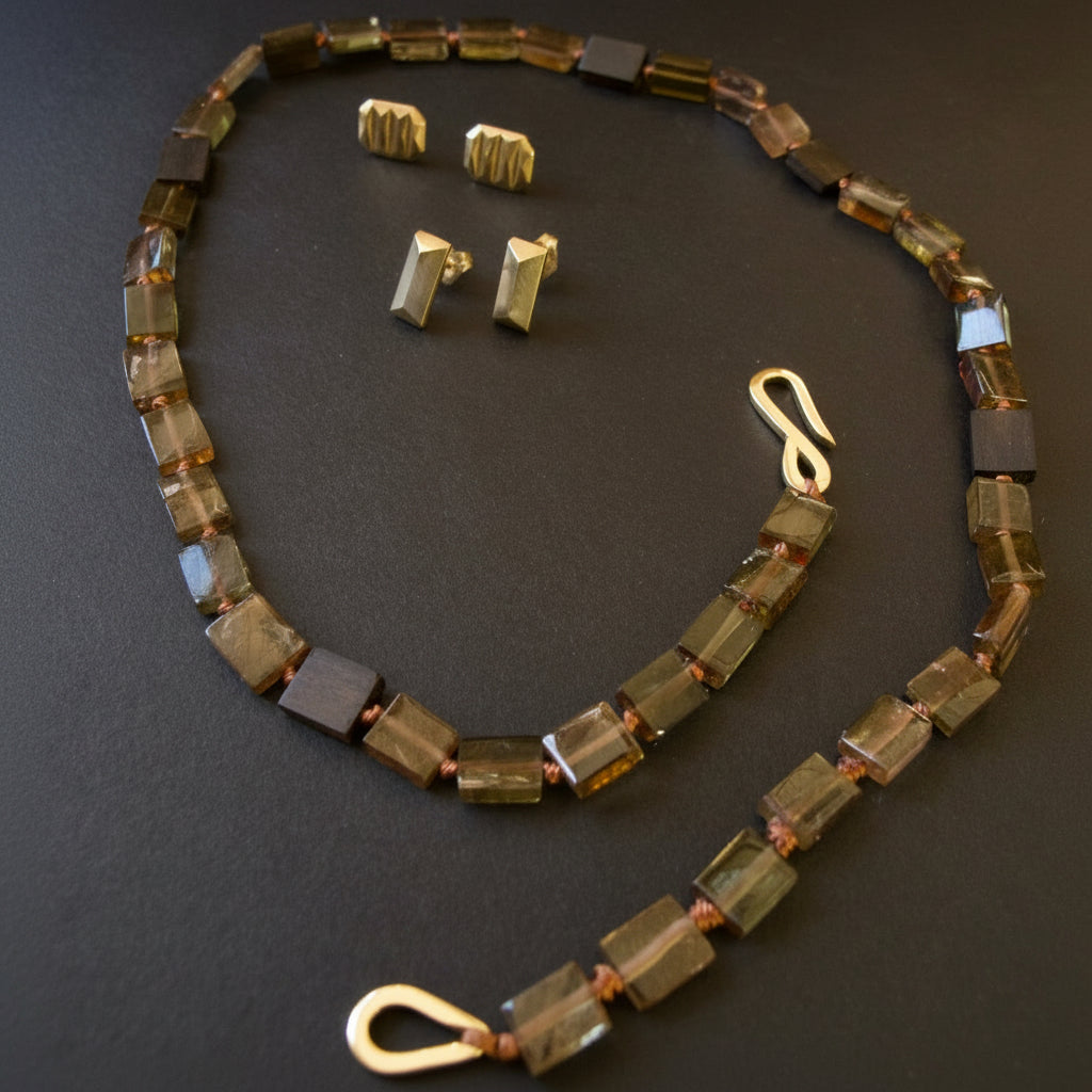 A beaded necklace and matching stud earrings laid out on a dark background. The jewelry features rectangular faceted beads in earthy brown, olive green, and smoky gray tones, accented with small gold-toned spacer beads and a gold lobster clasp.