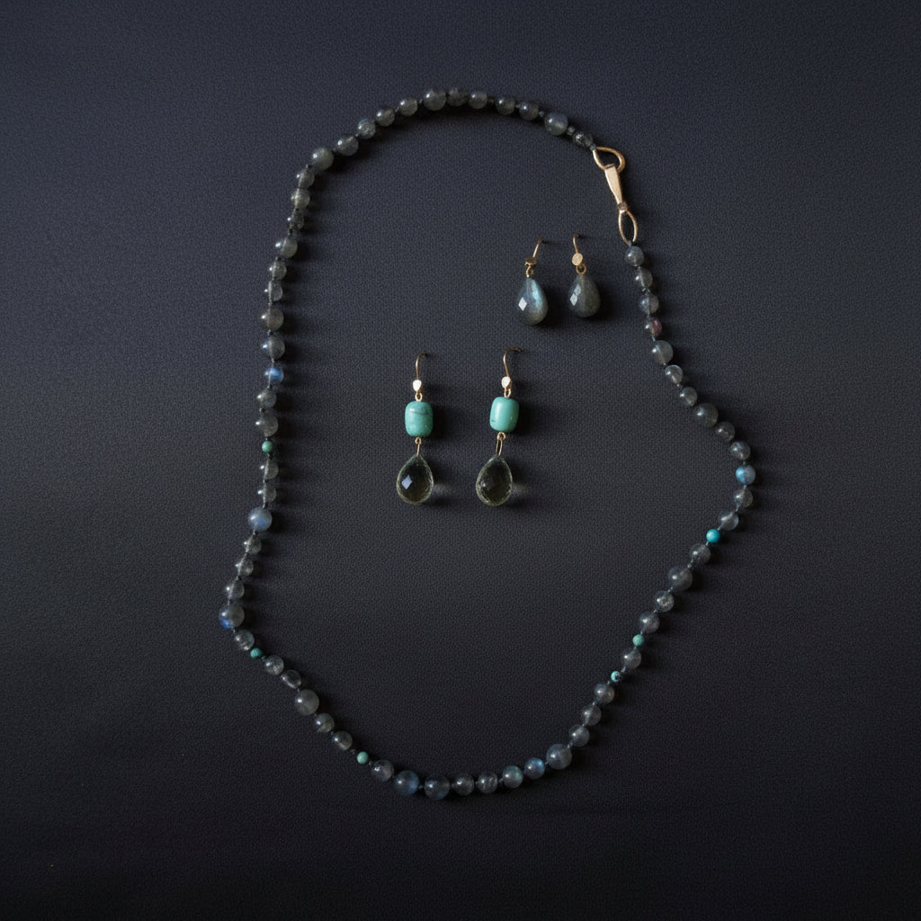 A jewelry set featuring a beaded necklace and matching drop earrings laid out on a dark satin fabric background. The necklace and earrings are made with small iridescent gray-blue beads, with one earring featuring a light turquoise bead and another with a dark teardrop-shaped stone.