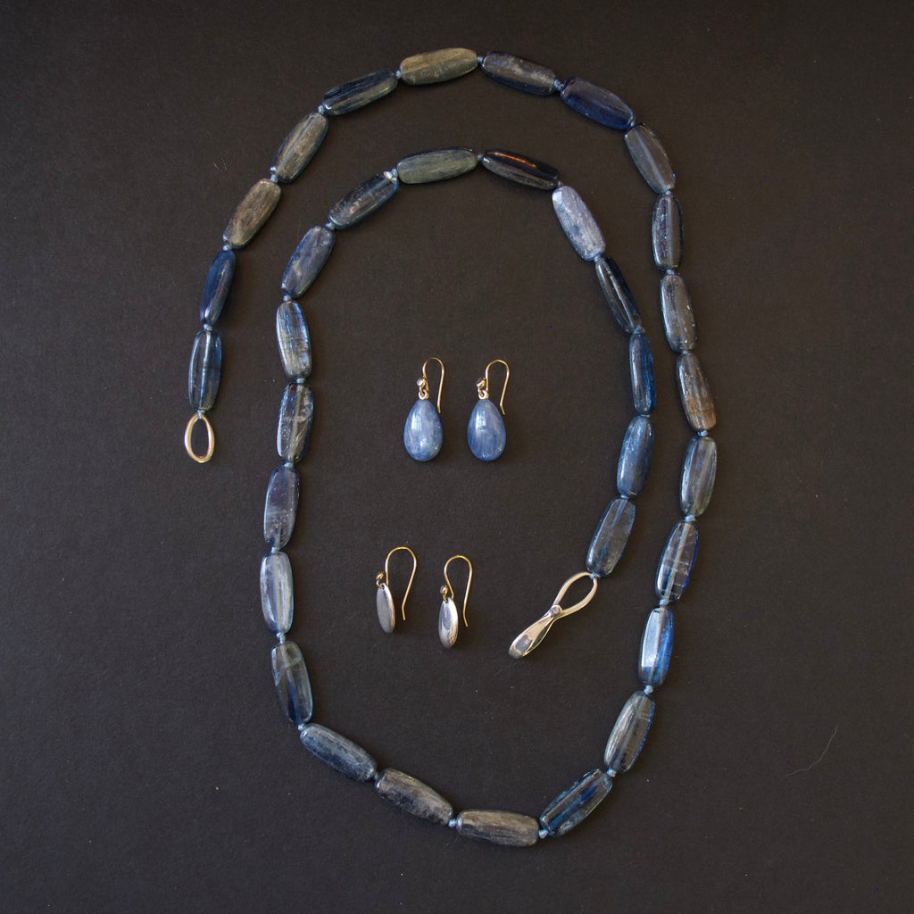 A jewelry set featuring a beaded necklace, matching drop earrings, and small stud earrings with blue-gray beads and gold-tone metal findings, arranged on a dark background.