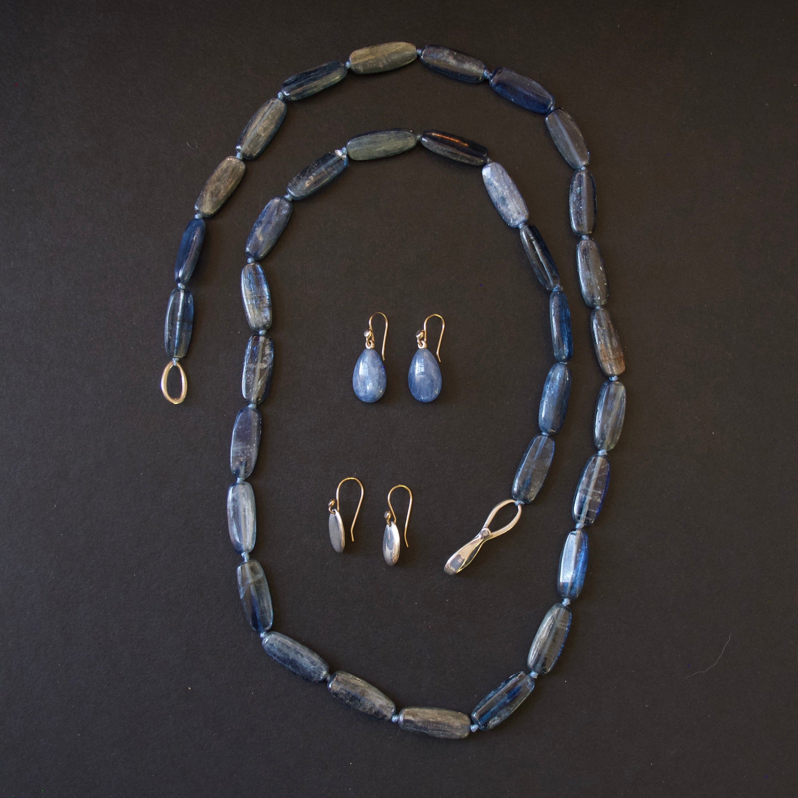 A jewelry set featuring a beaded necklace, matching drop earrings, and small stud earrings with blue-gray beads and gold-tone metal findings, arranged on a dark background.