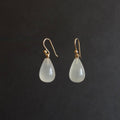 A pair of teardrop-shaped drop earrings with milky white translucent beads and gold fishhook wires, displayed against a dark gray background.