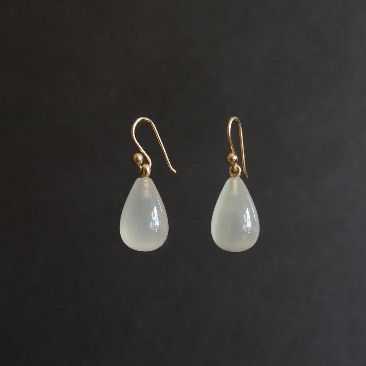 A pair of teardrop-shaped drop earrings with milky white translucent beads and gold fishhook wires, displayed against a dark gray background.